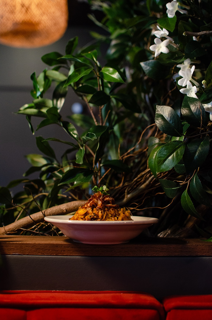 A bowl of kothu, or torn up bits of bread, is presented like a pasta dish and topped with a sprig of greenery, photographed in front of faux greenery with white flowers.