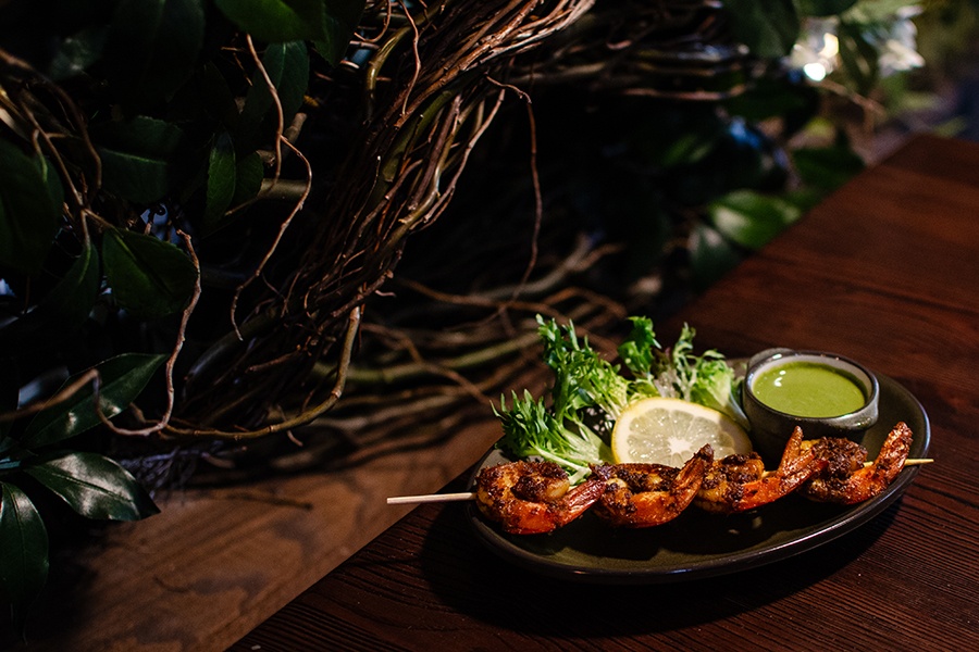 Four spiced shrimp on a skewer sit on a dark plate with a thick green sauce and a lemon slice.