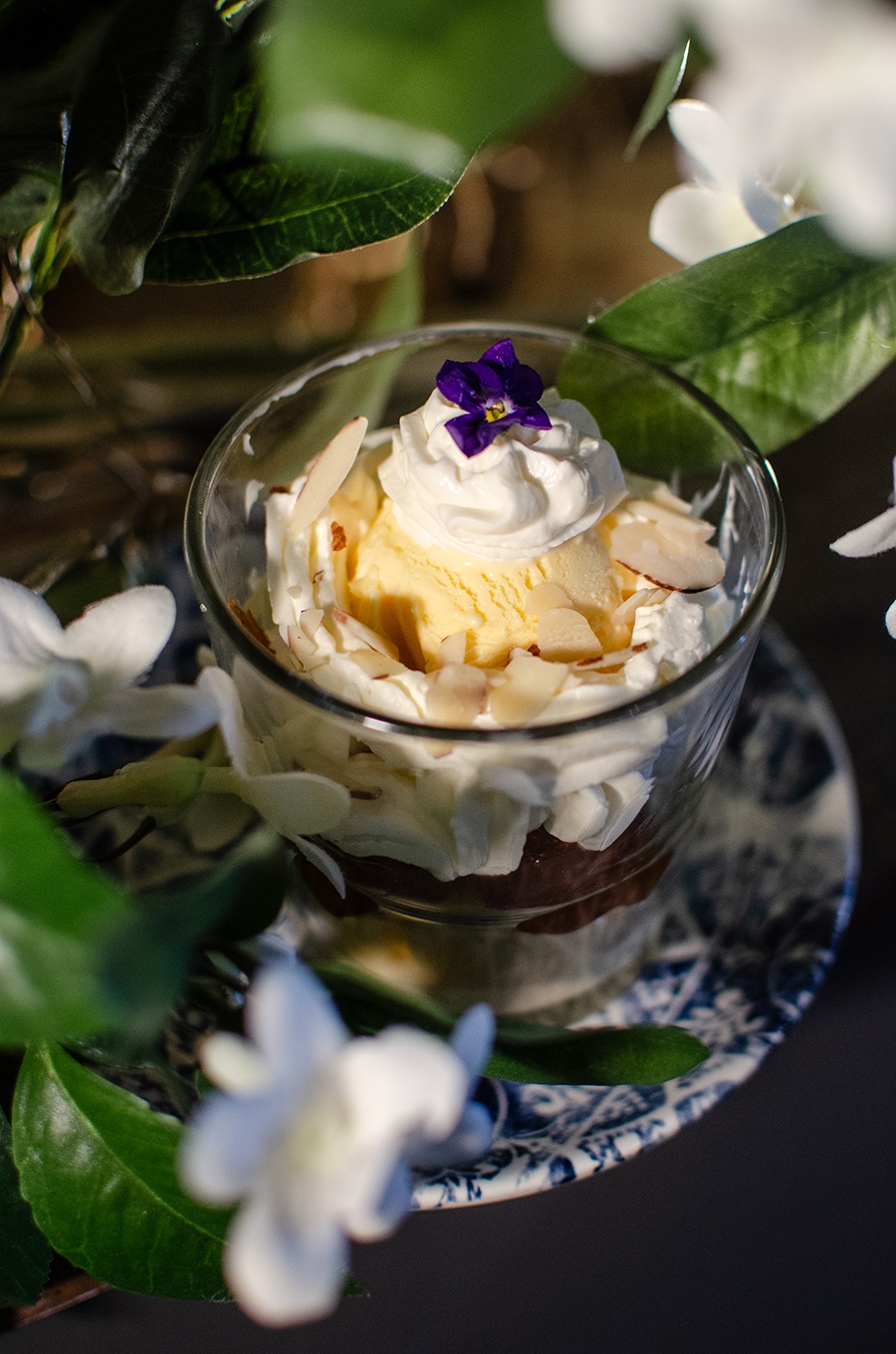 An ice cream sundae topped with whipped cream, slivered almonds, and a small purple flower is photographed among faux white flowers and green leaves.