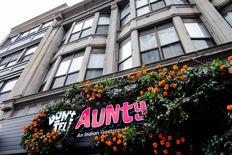 Exterior of a restaurant features signage in a bold font that says "Don't Tell Aunty: An Indian Gastropub." The signage is surrounded by green leaves and orange flowers.