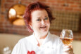 Chef Lydia Shire, with bright red hair and wearing chef's whites and a red dragon brooch, smiles at the camera while holding a glass of wine.