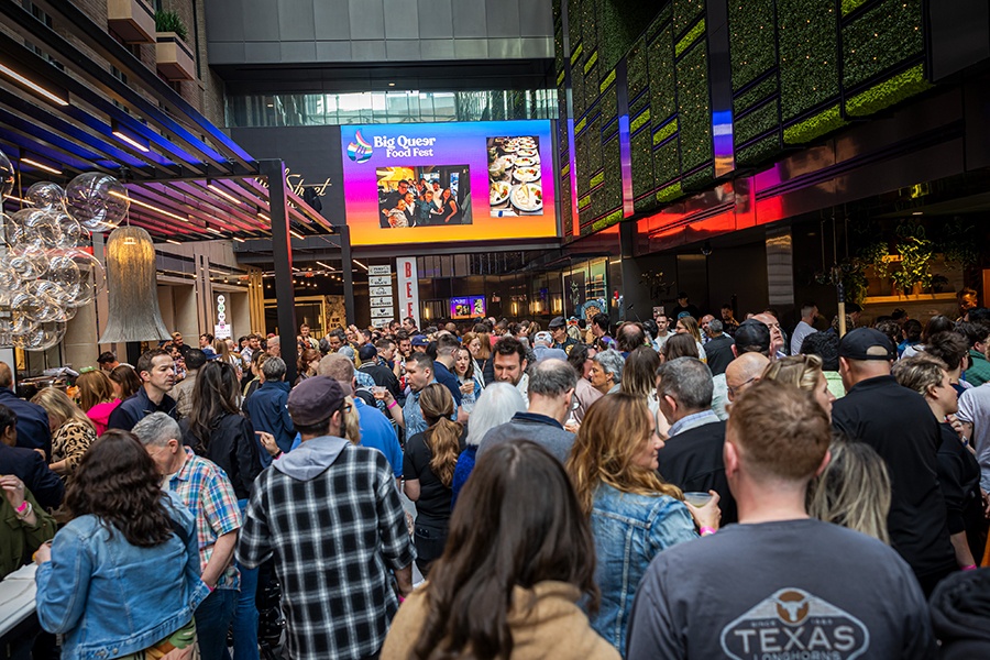 A crowd of people pack into a food hall with "Big Queer Food Fest" branding on a giant projection screen overhead.
