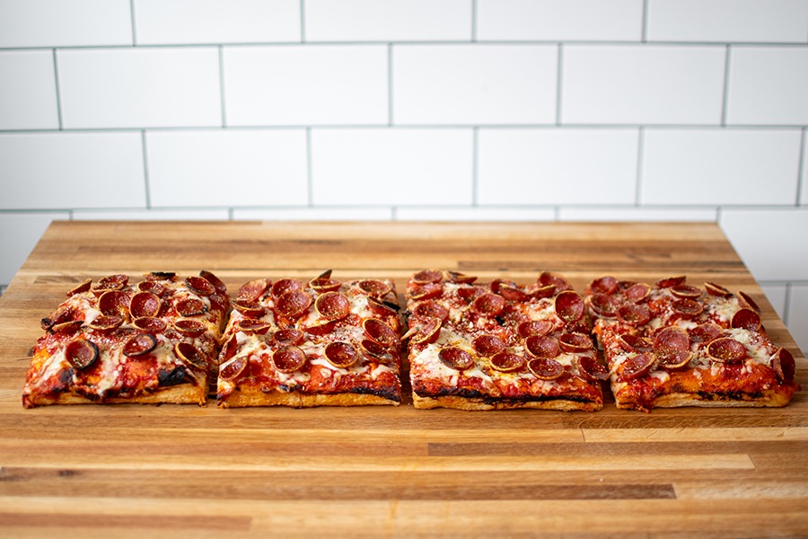 Four rectangular slices of Roman-style pepperoni pizza, with charred pepperoni cups, sit on a wooden counter in front of a white-tiled wall.
