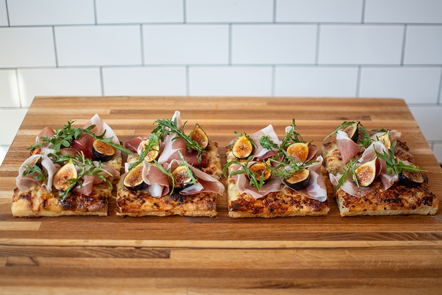 Four rectangular slices of Roman-style pizza, topped with half figs, sliced prosciutto, and arugula, sit on a wooden counter in front of a white-tiled wall.