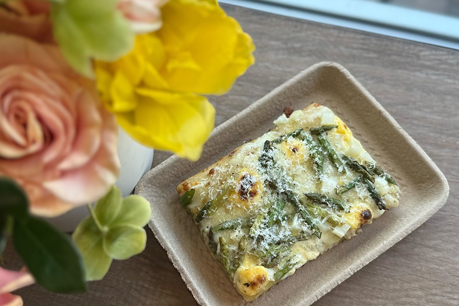A rectangular slice of white pizza with pieces of asparagus sits on a wooden counter next to a vase of flowers.