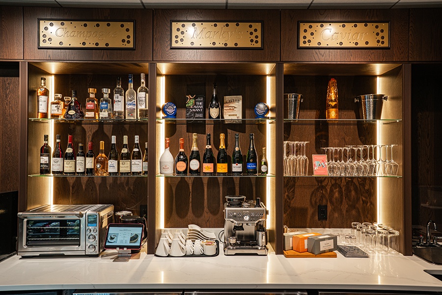 Glass shelves built into a dark wooden wall hold bottles of Champagne and liquor under golden signage that reads "Champagne" and "Marky's Caviar."