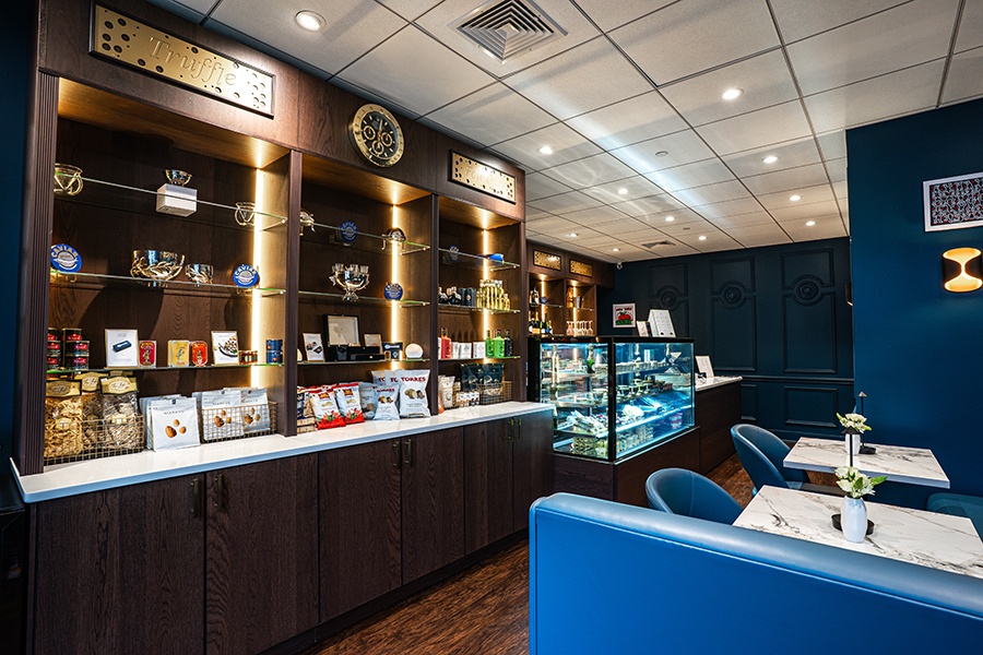A small retail shop and restaurant features shelves of truffles, foie gras, and other luxury food items, and a couple small white marble tables with royal blue chairs, banquettes, and walls.