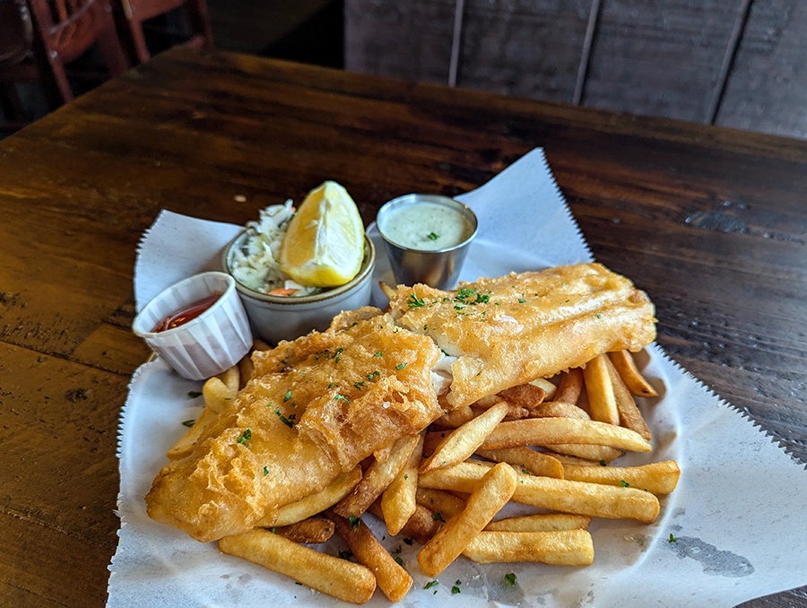 Big, crispy fish with fries, ketchup, coleslaw, a lemon wedge, and tartar sauce on a dark wooden table.