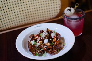 A plate of crispy glazed chicken with peanuts and daikon sits next to a pink cocktail with a black salt rim and lychee garnish.