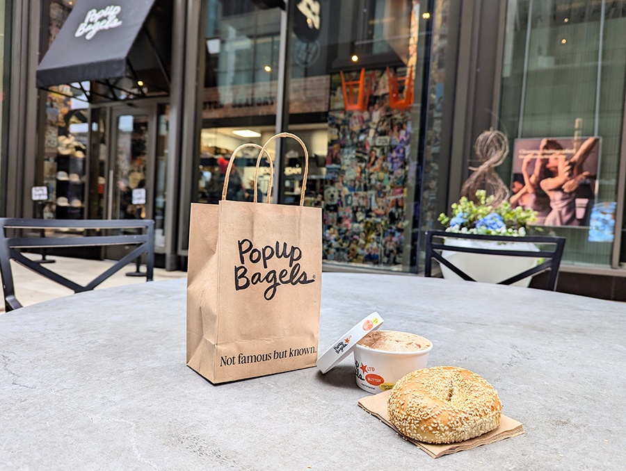 A brown bag with PopUp Bagels branding and a sesame bagel and butter container sit on an outdoor table outside of a bagel shop.
