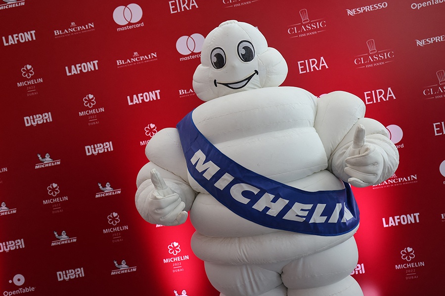 A person in an inflatable Michelin Man suit stands in front of a red backdrop covered with brand names at an event, giving two thumbs up.