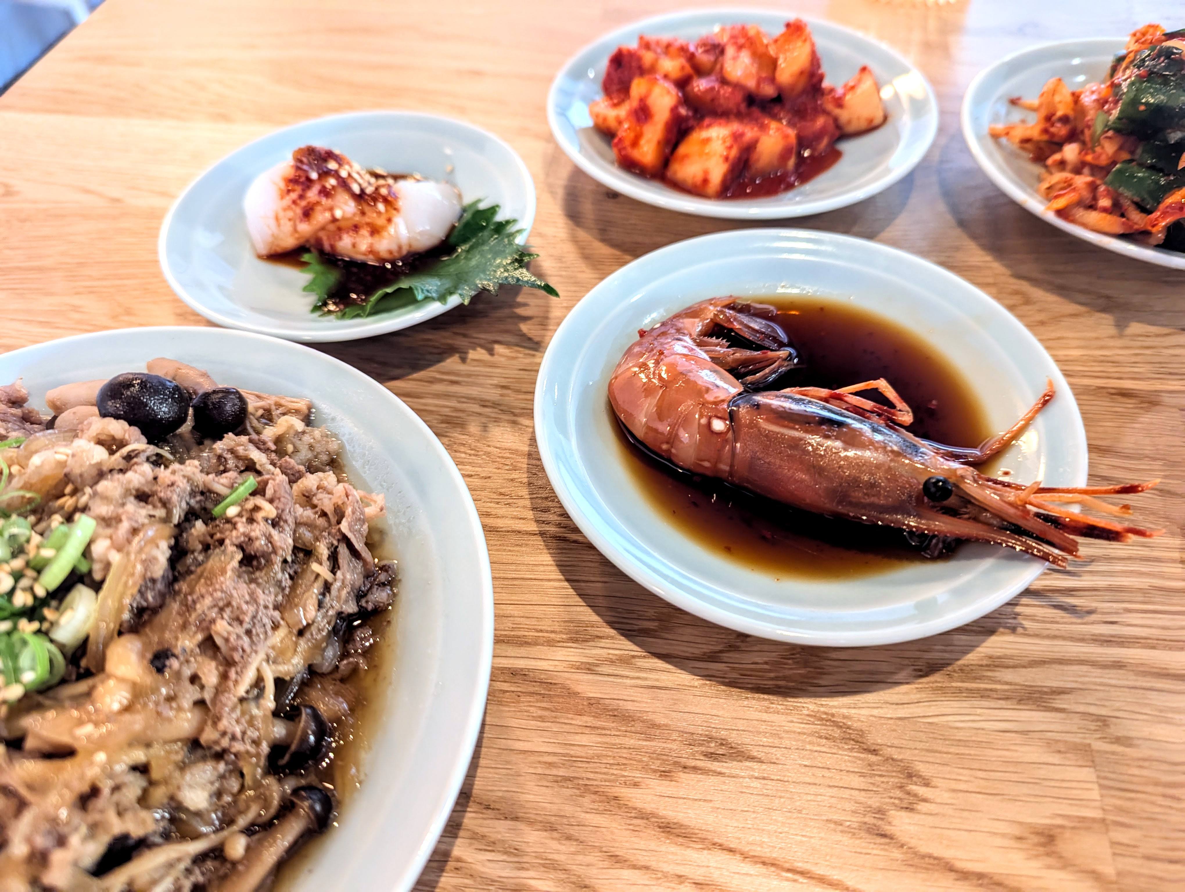 Small Korean plates on a light wooden table, including bulgogi, a big raw shrimp in a pool of soy sauce, raw scallops in a chili sauce, and radish kimchi.