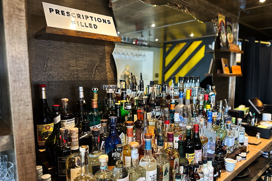 Shelves of liquor bottles on a backbar, decorated with signage that says "Prescriptions filled."