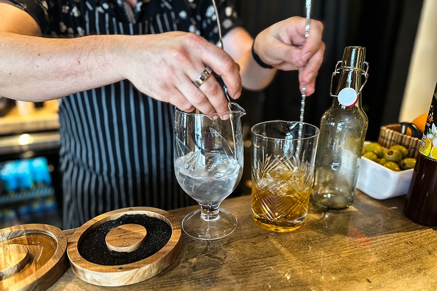 A bartender mixes two drinks with long silver stirrers.