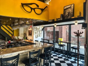 Interior of a bar with bright yellow walls, black-and-white checkered floors, and a big pair of decorative eyeglasses.
