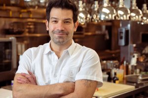 A chef in a white collared shirt stands, arms crossed, smiling at the camera in front of a restaurant kitchen.