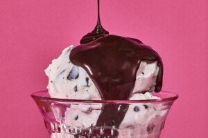 Hot fudge is poured over a scoop of white ice cream with chocolate chips in a glass bowl, photographed on a pink background.