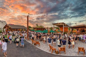 At sunset, lots of people gather at an outdoor dining, drinking, and live-entertainment venue with wooden Adirondack chairs, a stage, multiple bars, and some turf.