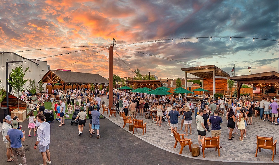 At sunset, lots of people gather at an outdoor dining, drinking, and live-entertainment venue with wooden Adirondack chairs, a stage, multiple bars, and some turf.