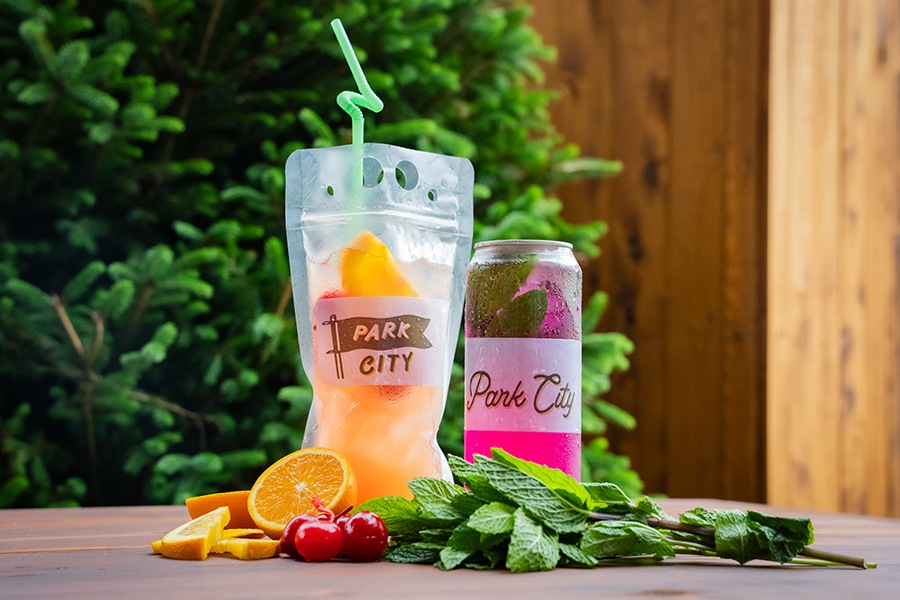 Two cocktails, one in a plastic pouch with bendy straw and one in a plastic can, are displayed on a wooden table in front of a tree.