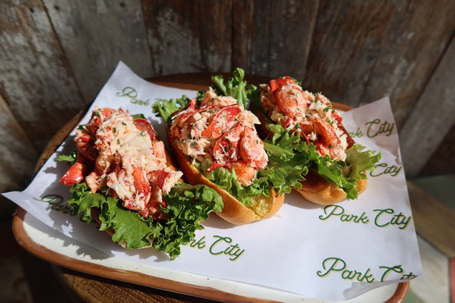 Three lobster rolls with mayo and lettuce are served in hot dog buns, sitting on a piece of paper that says Park City all over it.