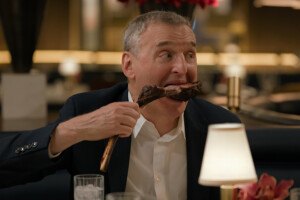 A man with gray hair and a suit coat over a collared, button-down shirt sits in an upscale restaurant and takes a big bite out of a piece of meat on a bone.