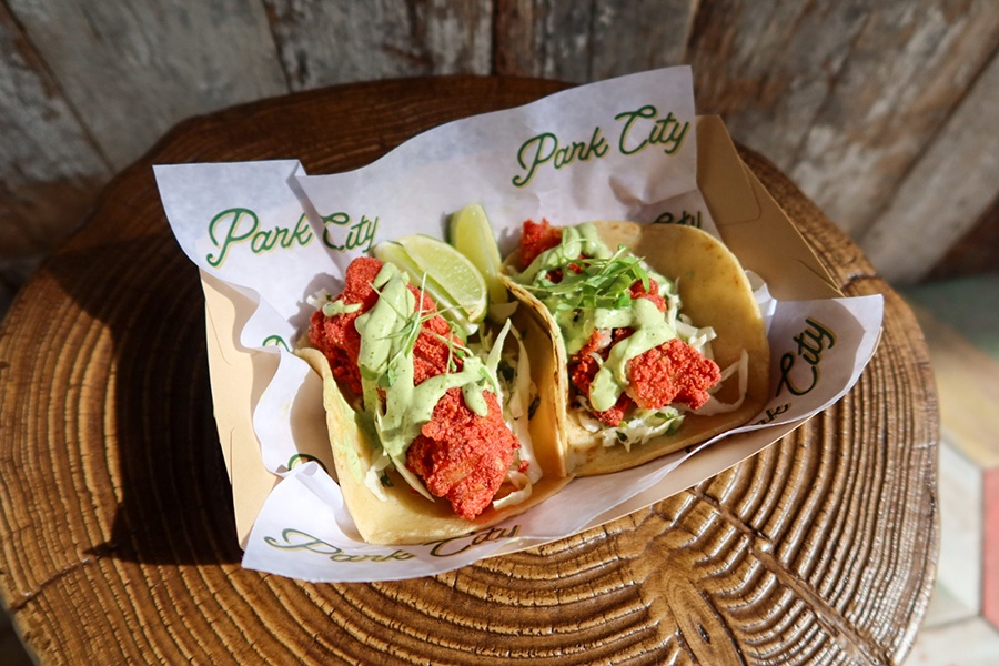 Fried fish tacos with avocado crema sit on paper that says Park City in a basket on a tree stump.