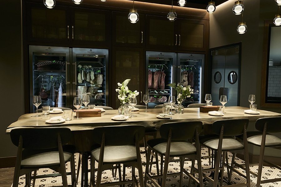 A communal dining table inside of a restaurant is positioned in front of dry-aging fridges full of meat.