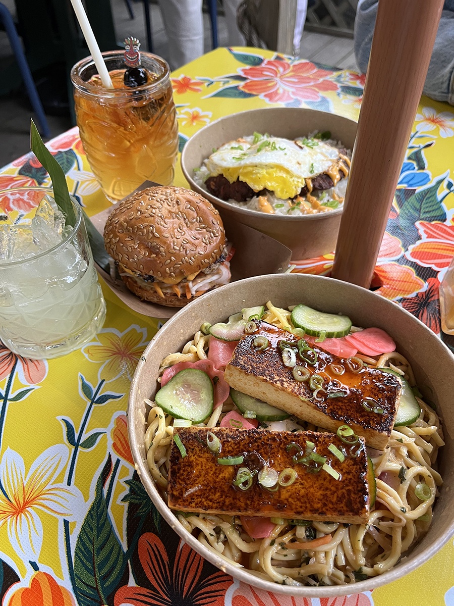 Salmon over noodles, a burger, and tiki drinks sit on a floral-patterned table.