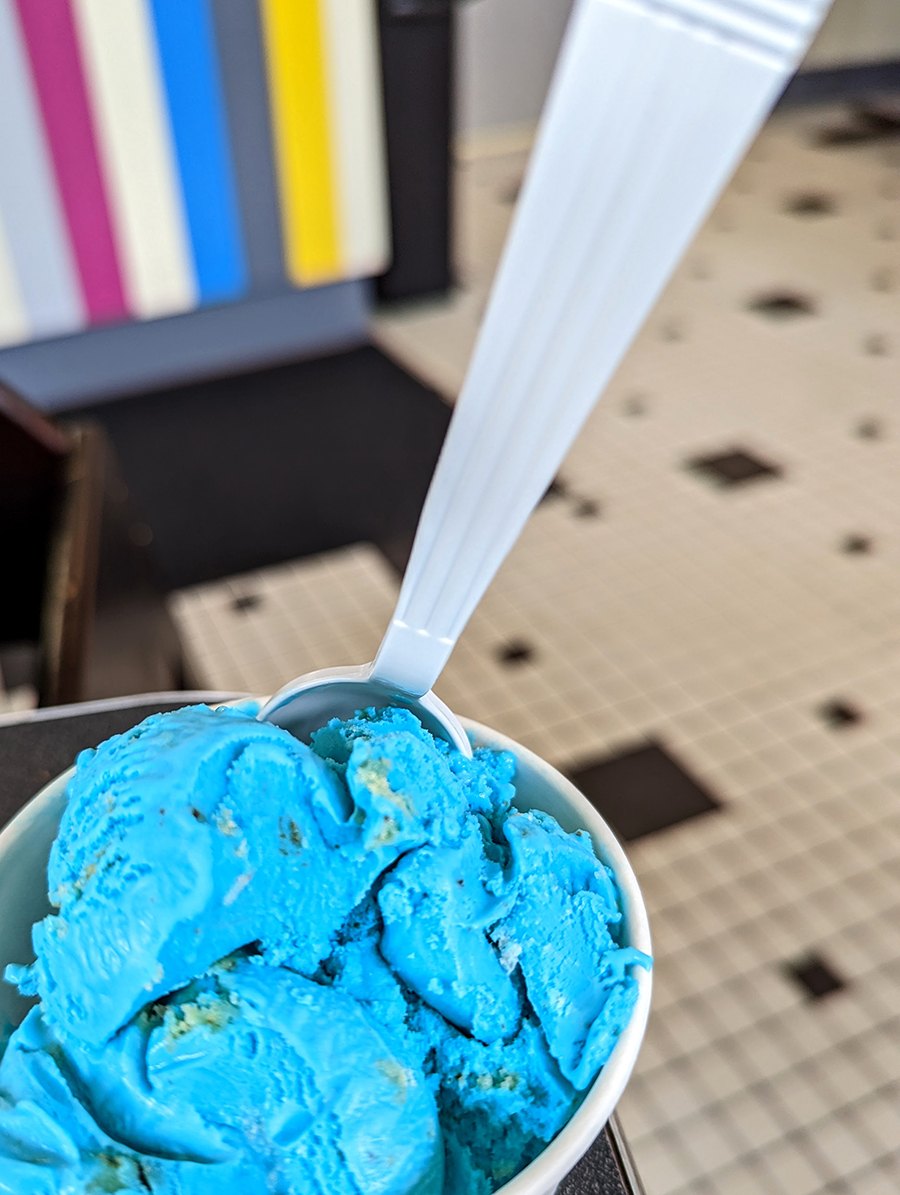 Bright blue ice cream in a cup is photographed with a colorful striped counter and a checkered tile floor in the background.