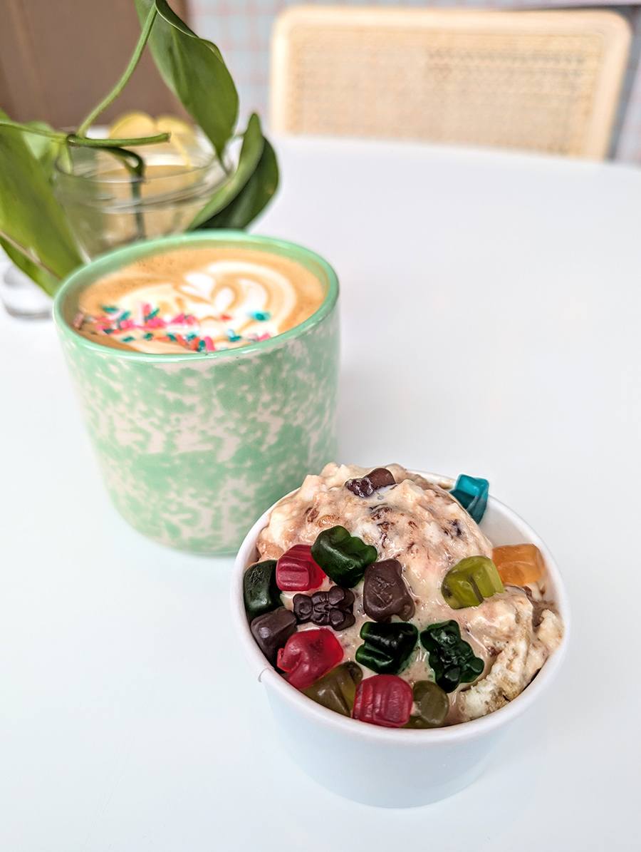 A cup of gummy bear-topped ice cream sits on a white table next to a green and white mug of a rainbow sprinkles-topped latte.