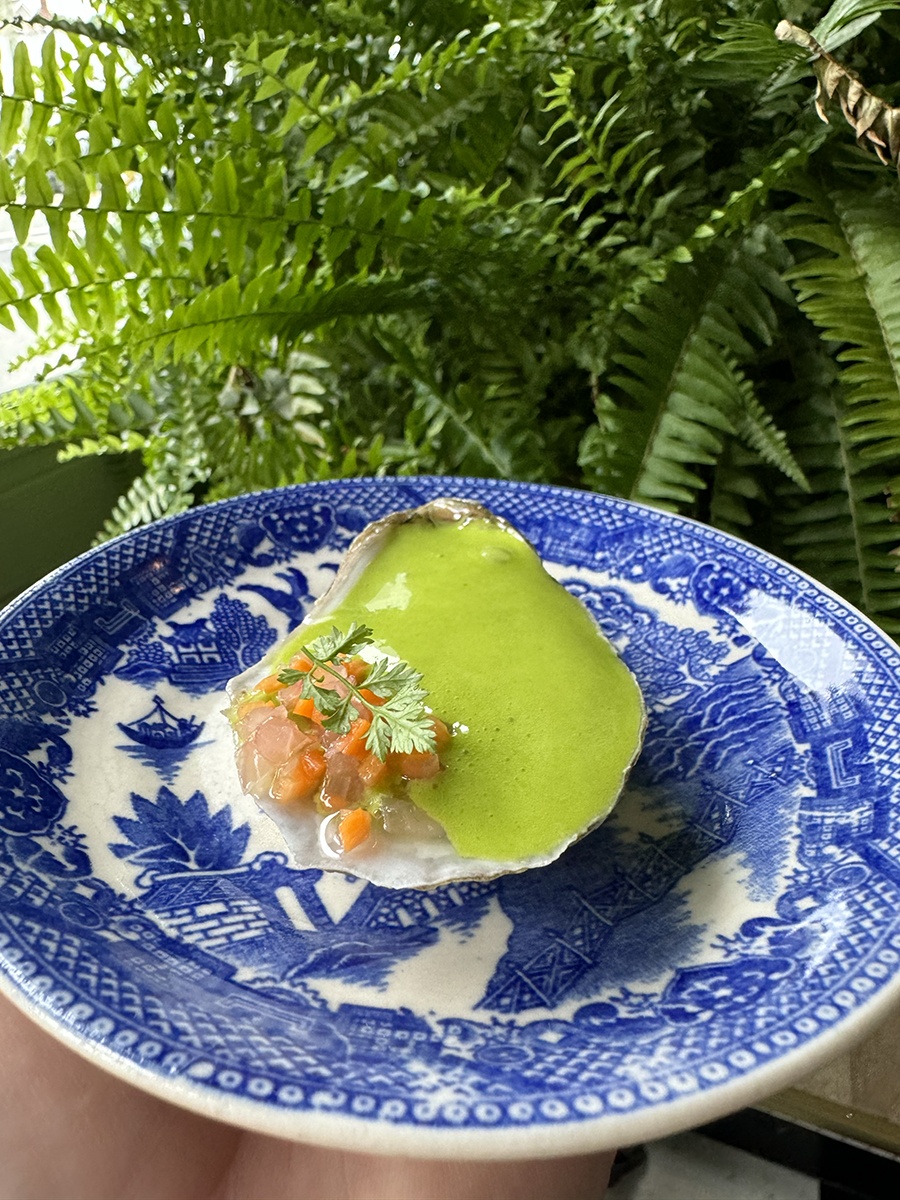 An oyster topped with green foam sits on a vintage-looking blue and white plate in front of ferns.
