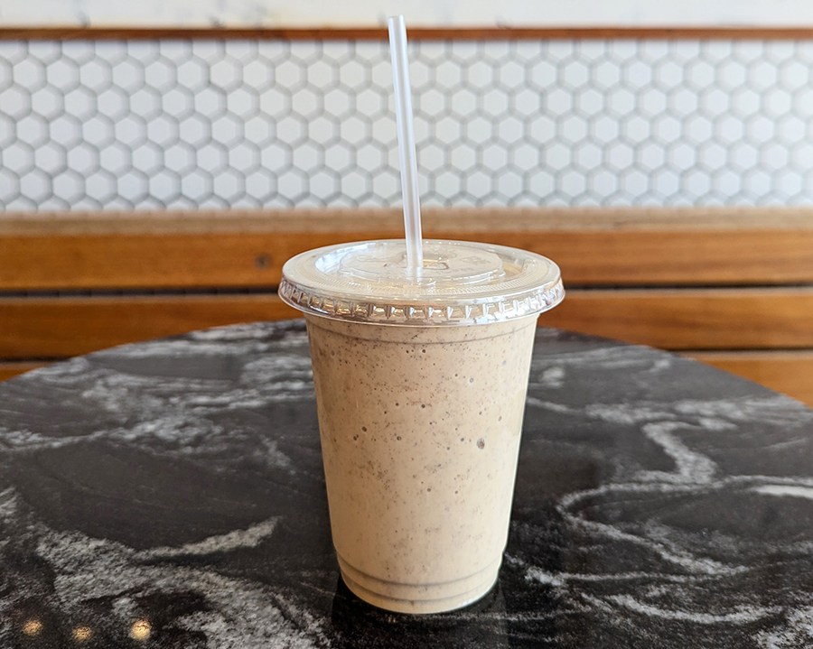 A pale brown milkshake in a plastic cup sits on a black marble table in front of a white tiled wall.