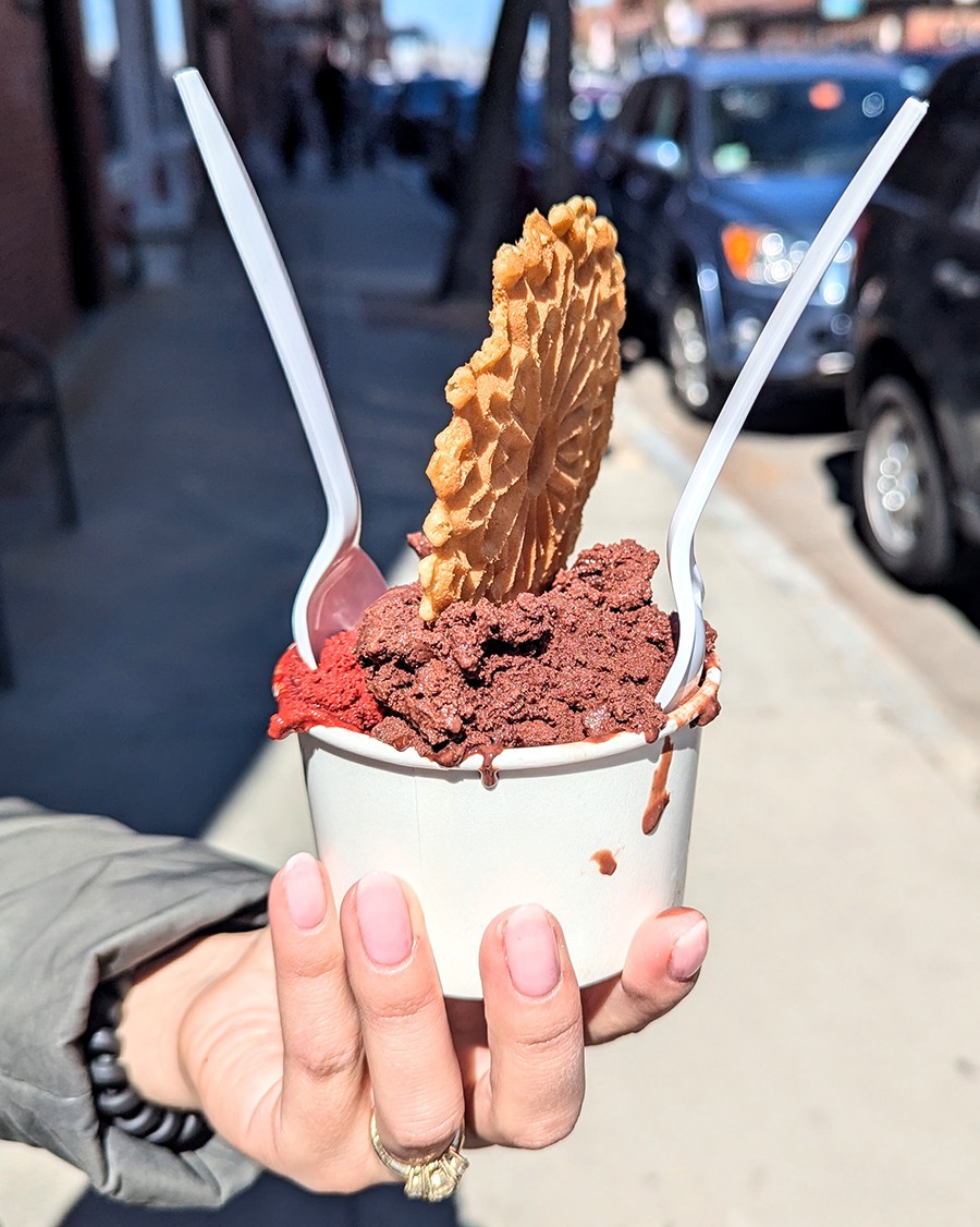 A hand holds a cup with two flavors of gelato (one red, one brown), garnished with a cookie, on a city street.
