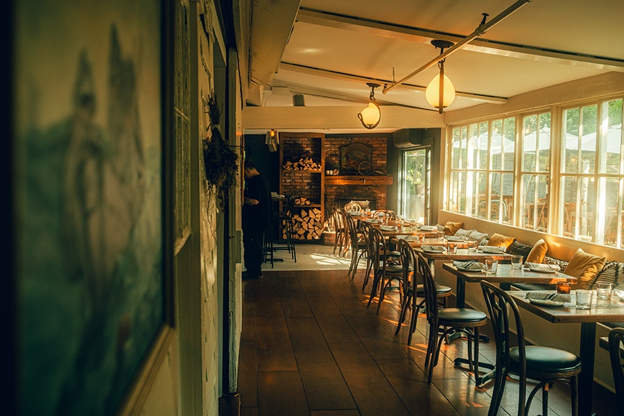 An empty restaurant dining room, long and narrow, is photographed with golden dusk light coming through the windows, and there's a brick fireplace at one end.