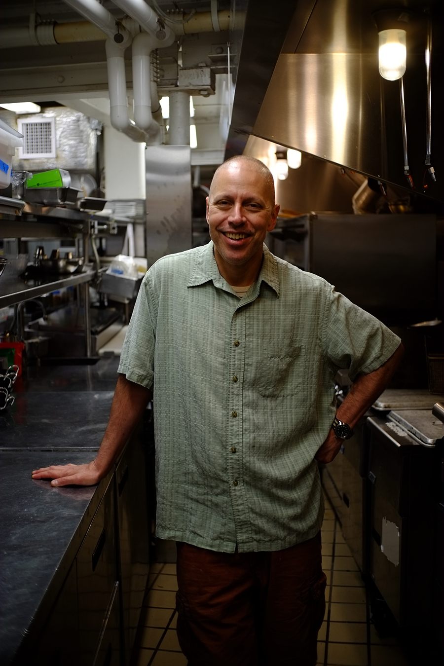 A man in a pale green collared shirt stands in a restaurant kitchen, hand on the counter, smiling at the camera.