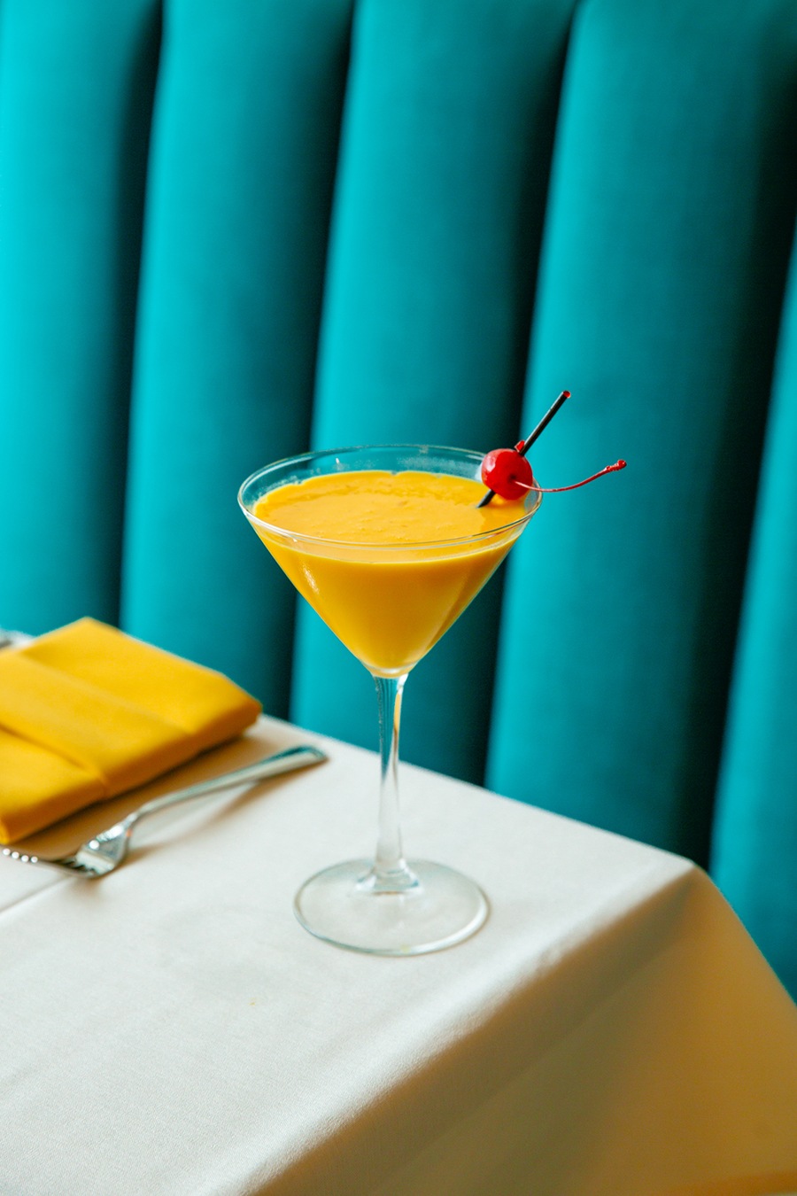 A thick orange cocktail garnished with a cherry is served in a martini glass in front of a teal banquette.