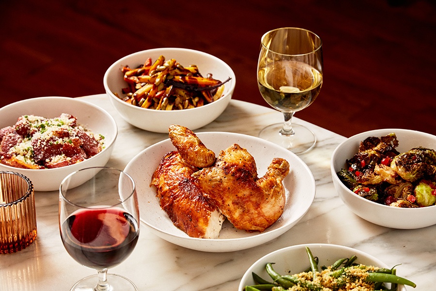Roast chicken, Brussels sprouts, crispy potatoes, and other sides are served on a white marble table with glasses of wine.