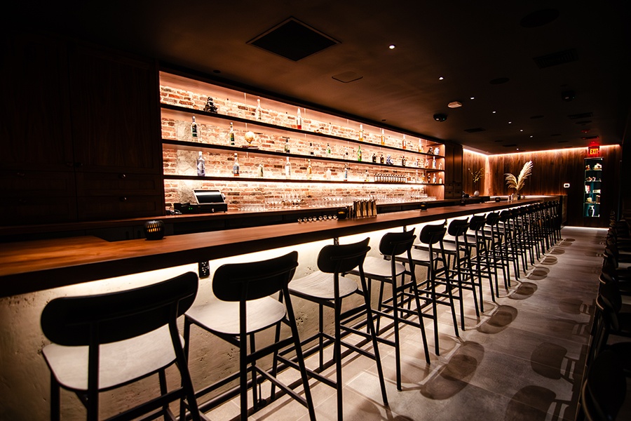 A long, narrow walnut bar is dimly lit. There's exposed brick behind the back bar.