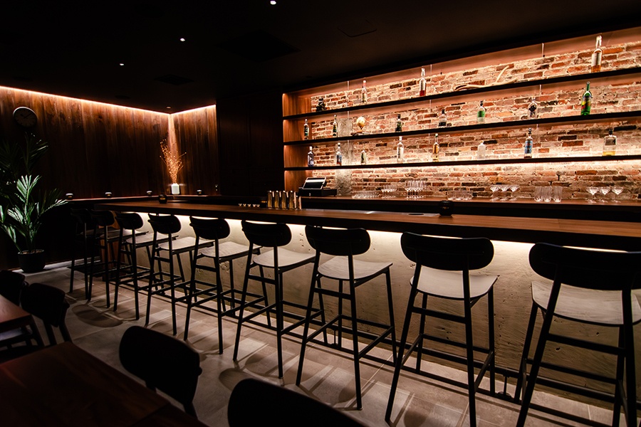 A long, narrow walnut bar is dimly lit. There's exposed brick behind the back bar, wood paneling on the other walls, and a couple plants decorating the space.