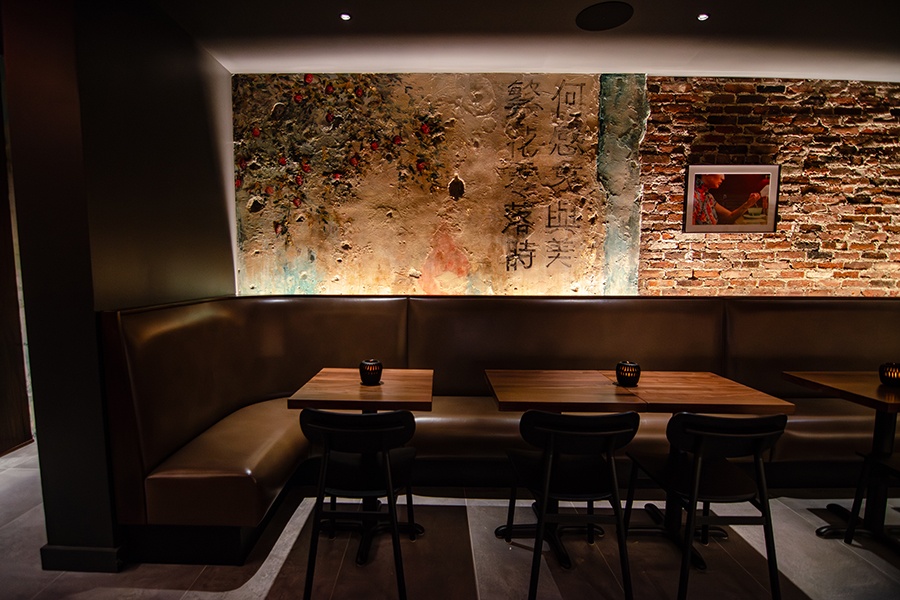 A restaurant interior features exposed brick, an aged-looking mural of a tree and Cantonese characters, and a dark brown banquette.