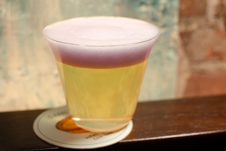 A clear yellow cocktail is topped with a thick pale purple foam, giving it a beer-like appearance.