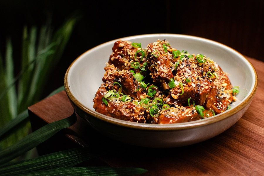 Ribs in a brown glaze, garnished with sesame seeds and scallions, sit in a bowl on a wood counter.