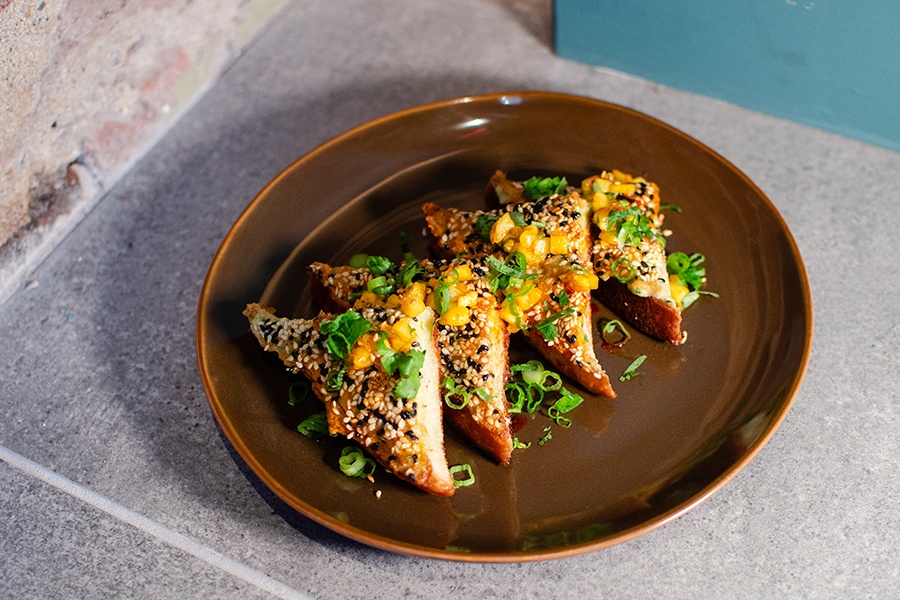 Four triangles of sesame toast are garnished generously with black and white sesame seeds.