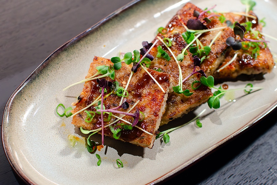 Four squares of turnip cake sit on an oblong plate, garnished with little green sprouts and a brown sauce.