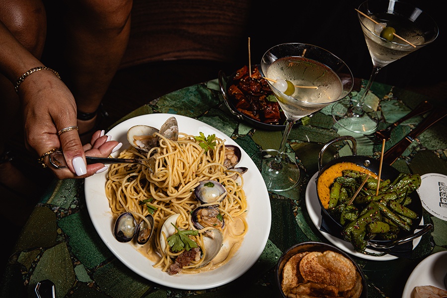 A hand picks up a forkful of linguine, tossed with clams, served on a table with martinis and apps.