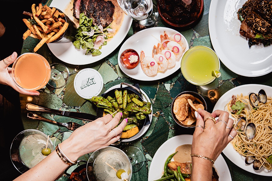 Hands pick up food and drink from a green table covered with linguine and clams, steak frites, shrimp cocktail, and more.