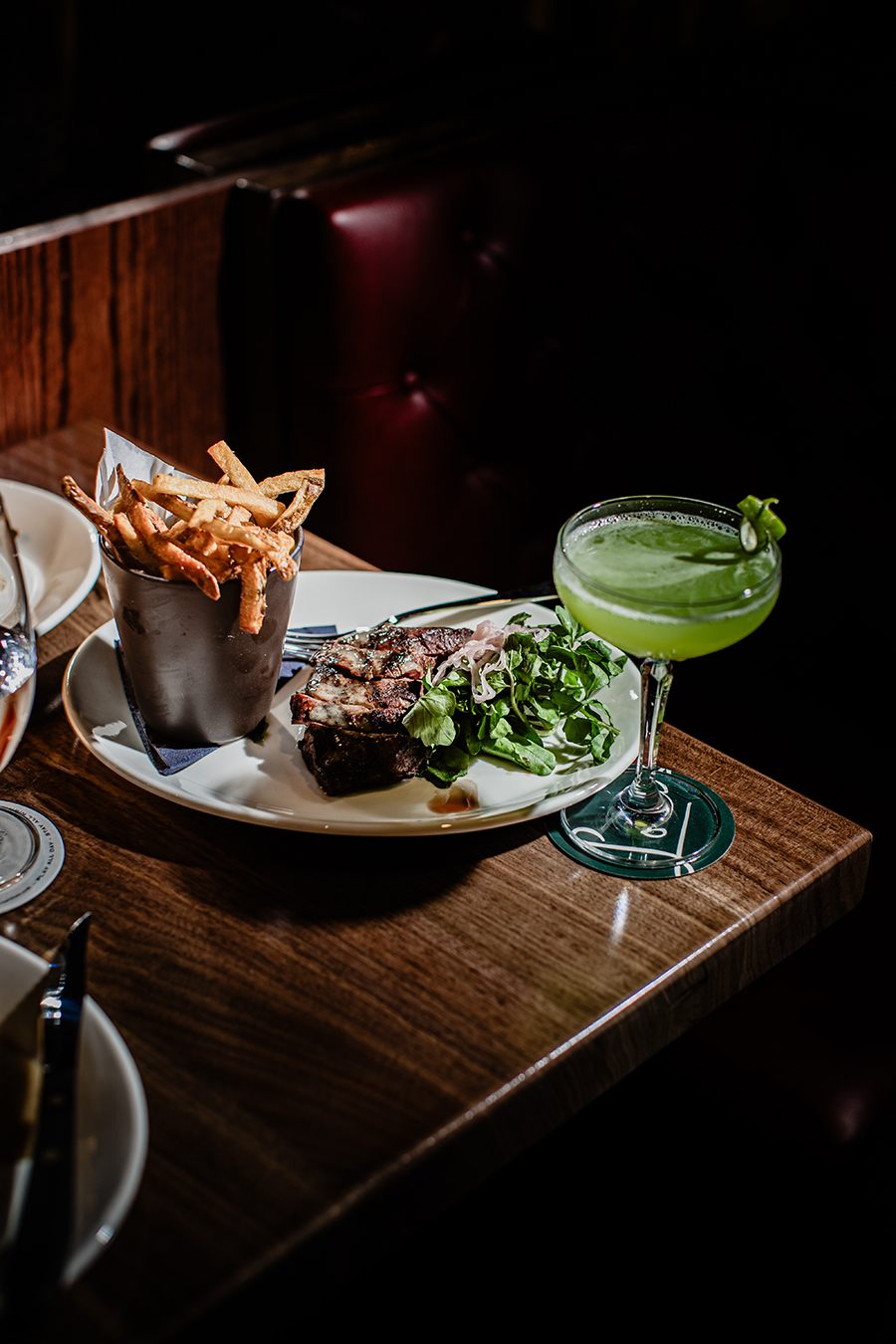 Steak frites and a green cocktail sit on a wooden table in a dark restaurant.