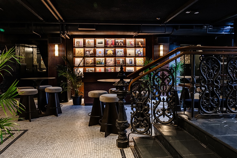 Stairs with an ornate black railing descend into a white-tiled, dimly lit restaurant space, featuring a wall of vinyl records.