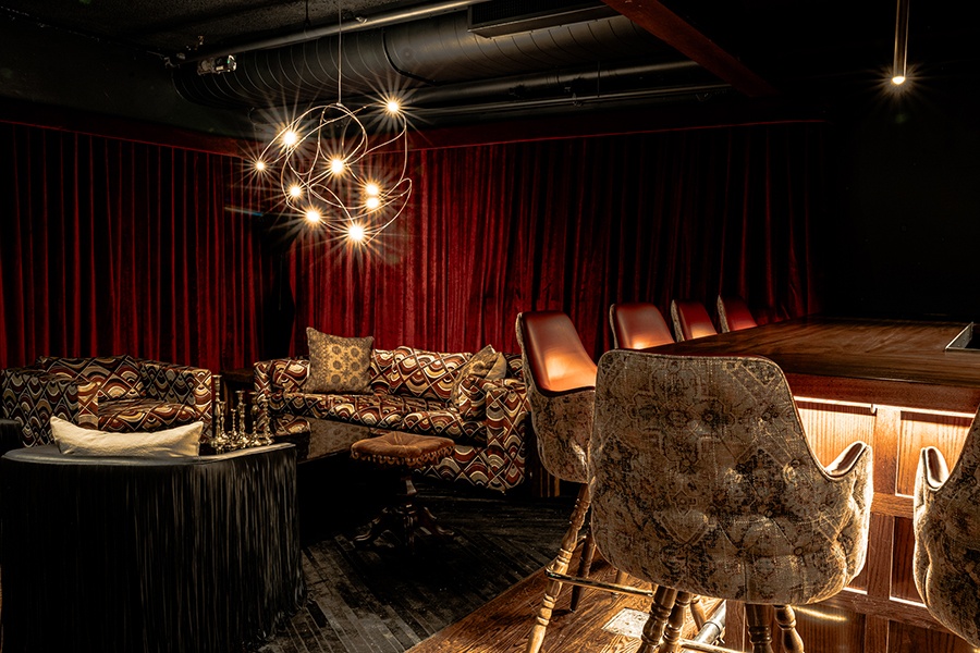 A dark bar and lounge has thick red velvet curtaining hanging in the background and couches and bar seats in mismatched upholstery.